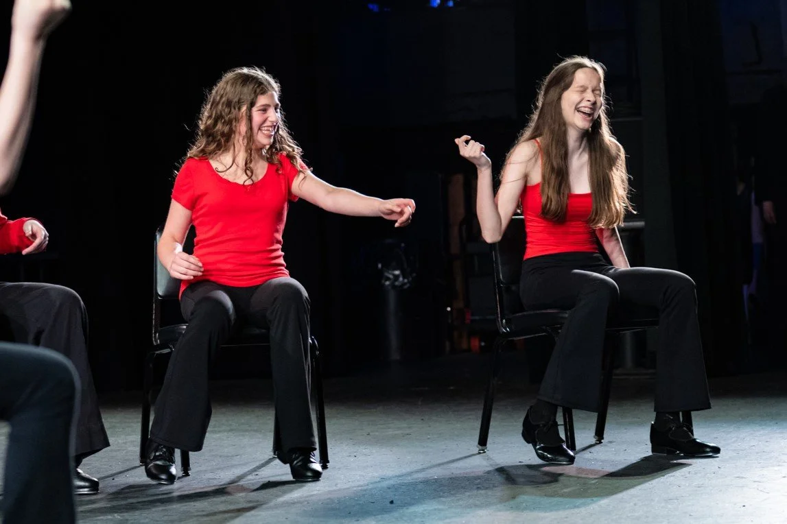 Two women with long hair sitting on chairs on a stage, laughing and joking with each other, wearing red tops and black pants.