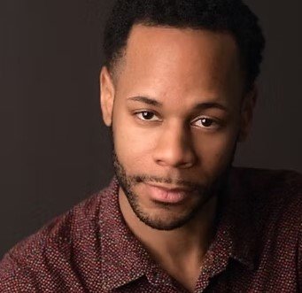 A young man with short curly hair and a beard, wearing a dark patterned shirt, looking at the camera with a slight smile against a dark background.