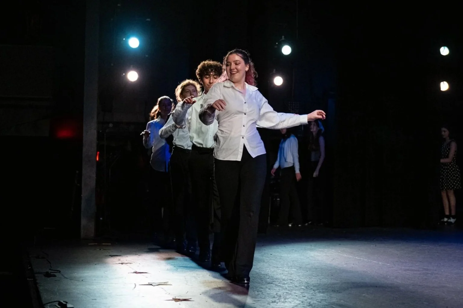 Group of women in white shirts and black pants performing a dance on stage with stage lights overhead.