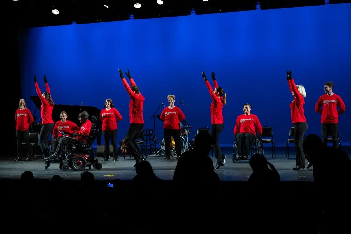Group of performers on stage, some in wheelchairs, wearing red sweatshirts, dancing and singing with blue backdrop and musical instruments.