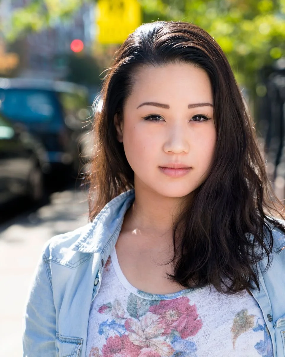Close-up of a young woman with long dark hair, wearing a floral shirt and denim jacket, standing outdoors on a sunny day with blurred trees and parked cars in the background.