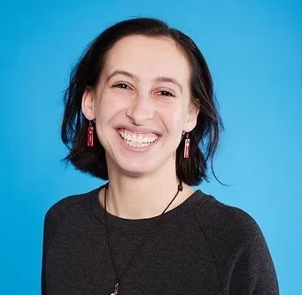 A woman with short dark hair, smiling, wearing earrings and a black top, against a blue background.
