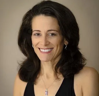 A woman with dark, shoulder-length hair smiling at the camera, wearing a black top and a necklace.