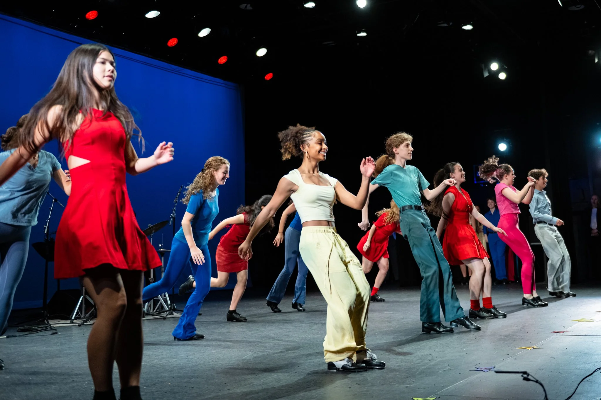 Group of young women dancing on stage with colorful outfits, black stage floor, blue background, and stage lights.