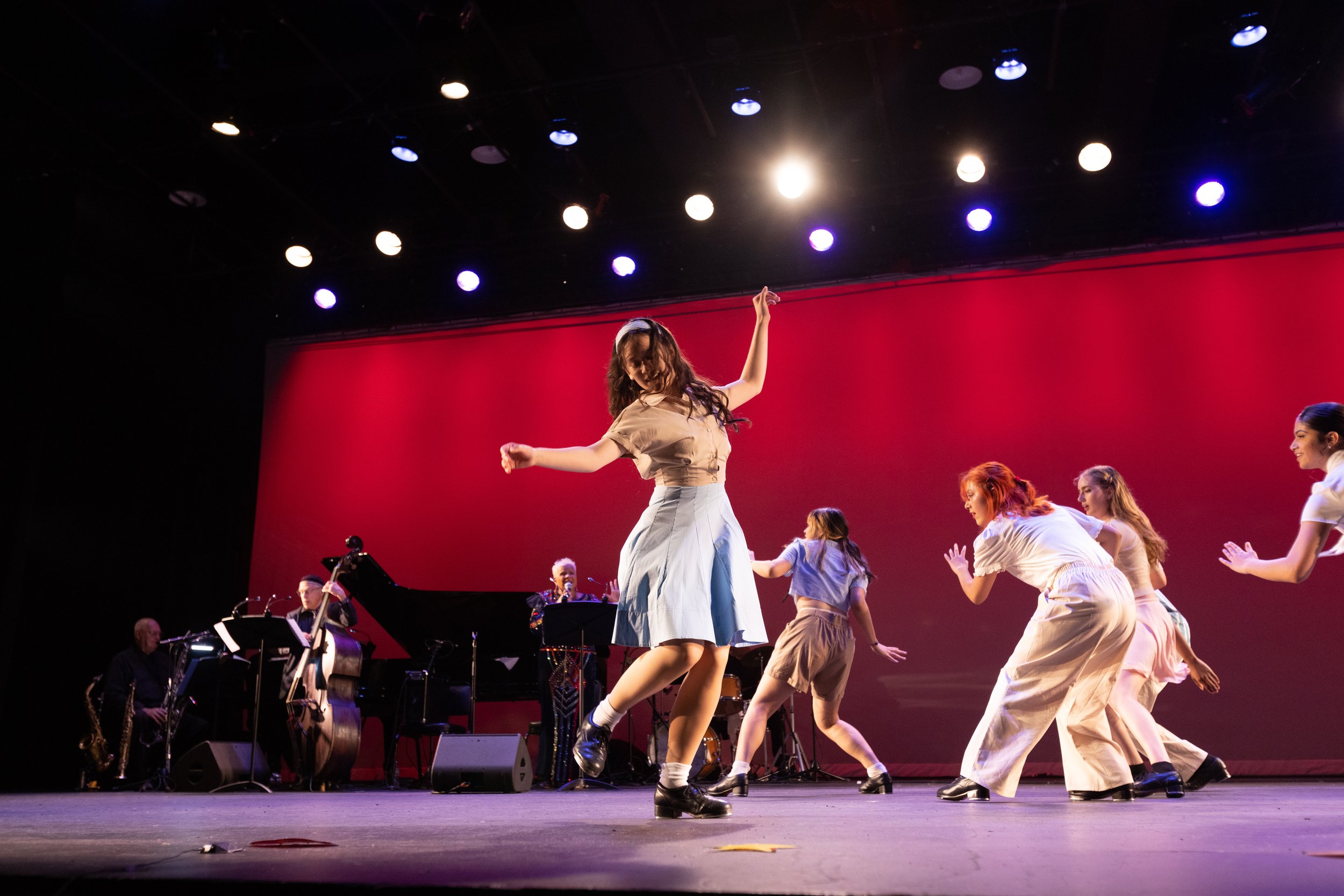Group of young women dancing on stage with a red background, accompanied by musicians and a pianist.