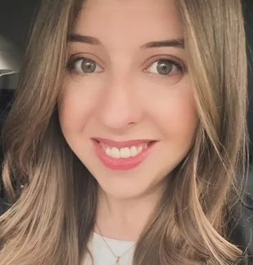 Close-up of a young woman with long, light brown hair, smiling showing her teeth, with fair skin and gray eyes.