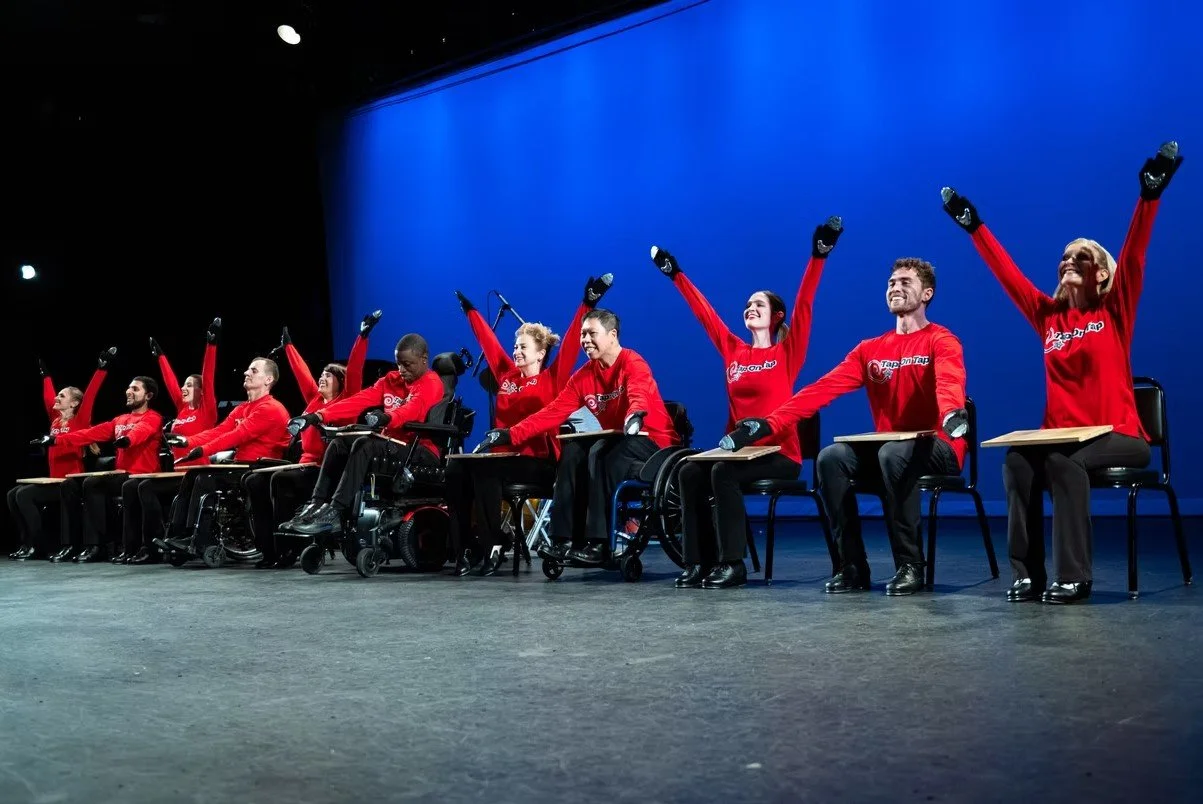 People with disabilities participating in a performance or event, wearing red shirts, sitting on chairs with some using wheelchairs, raising their hands in the air on a stage with a blue background.