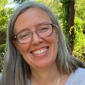 A smiling woman with gray hair and glasses outdoors in a forested area