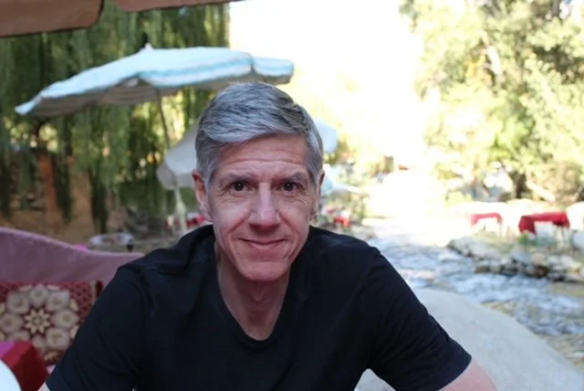 A man with gray hair smiling outdoors in front of a river with trees, a white umbrella, and outdoor seating.