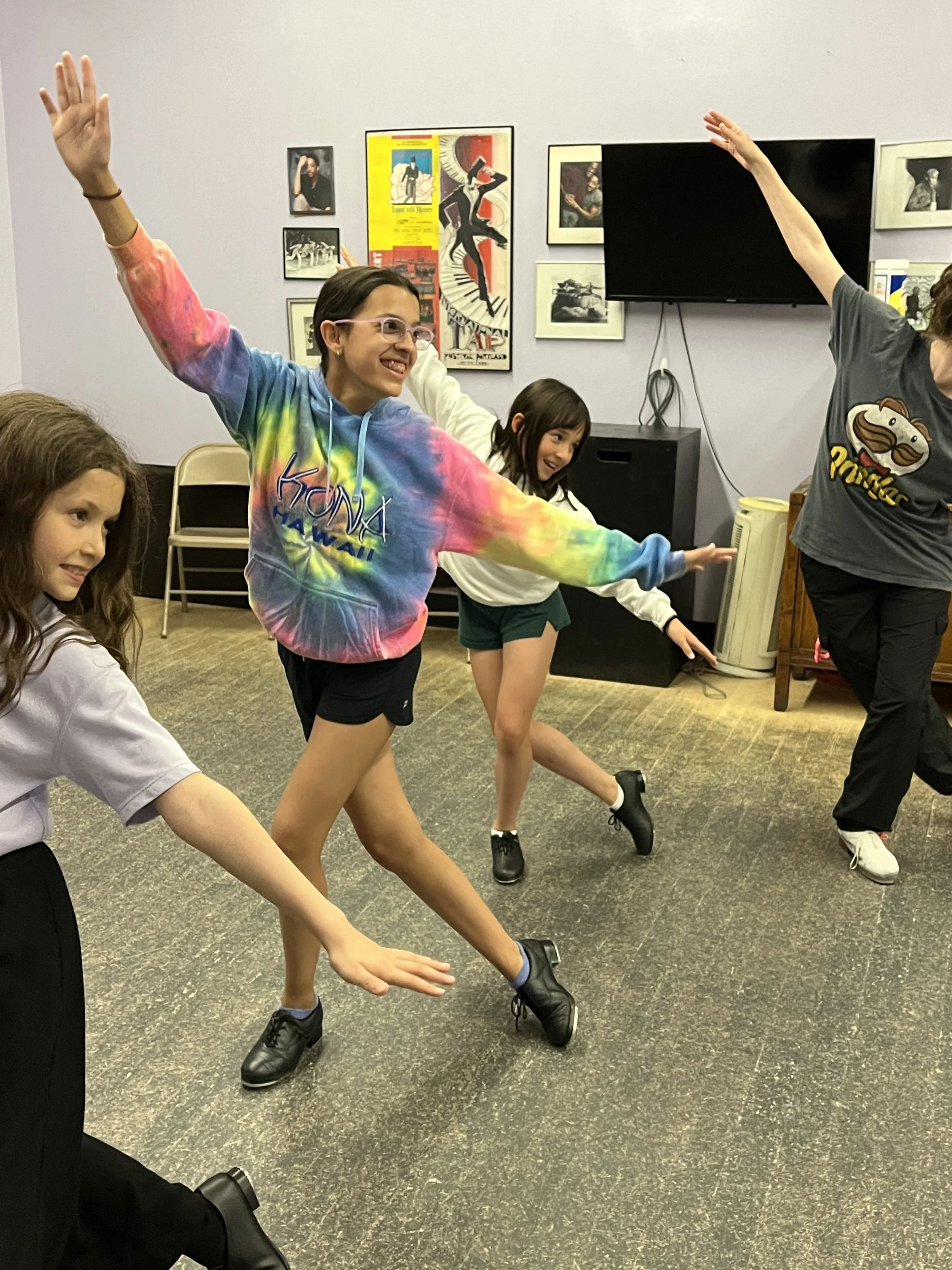 A group of young women dancing indoors with their arms outstretched, in a room with framed pictures and posters on the wall, a TV, and various furniture.