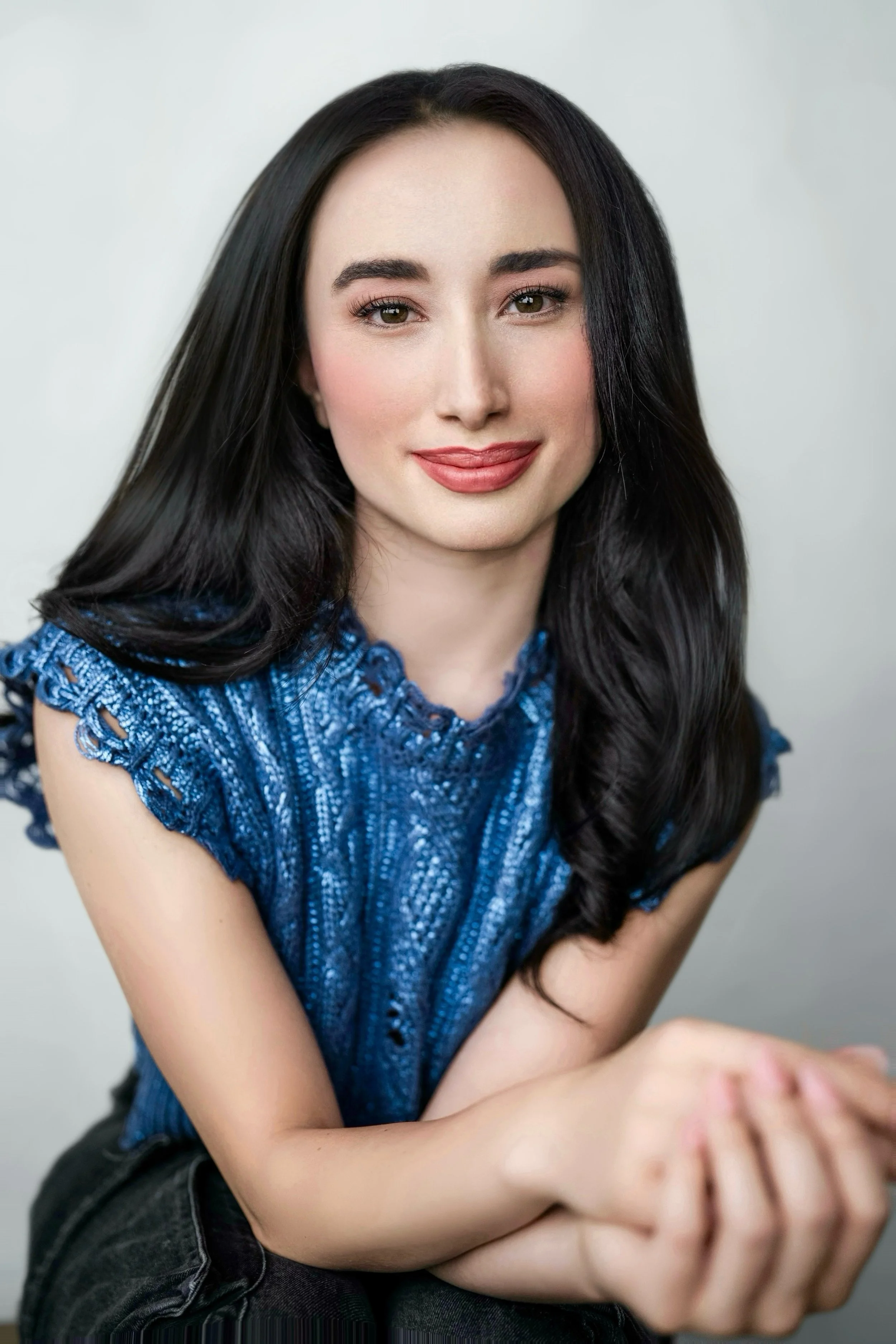 A woman with long dark hair, wearing a blue textured top, smiles softly with her hands clasped in front of her, in front of a plain light background.