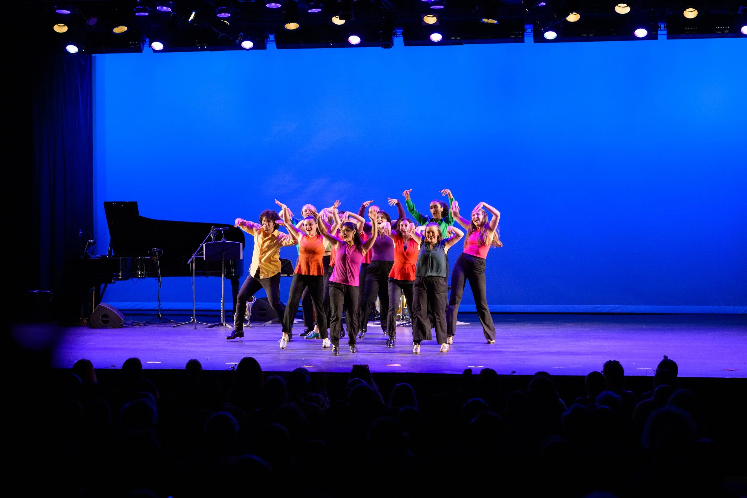 A group of dancers performing on stage with a blue backdrop, a piano on the side, and an audience watching.