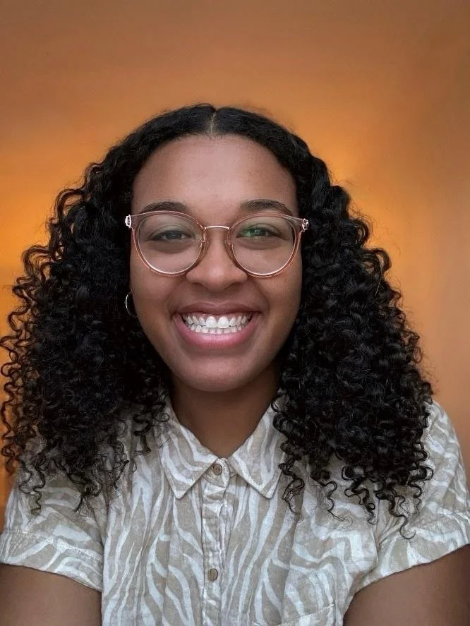 A young woman with curly black hair, wearing glasses and a light-colored patterned button-up shirt, smiling in front of an orange background.