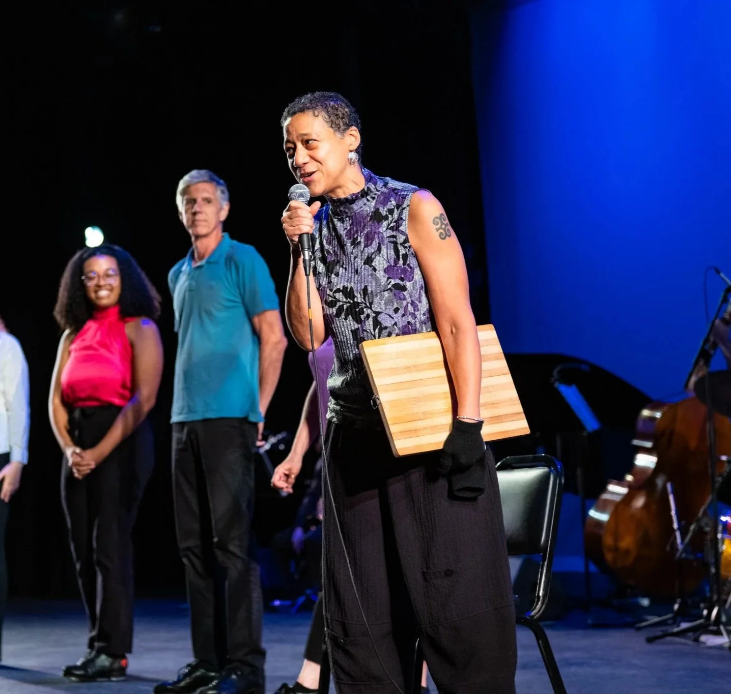 A woman speaking into a microphone on stage, holding a wooden clipboard, with three other people standing behind her. There are musical instruments in the background.
