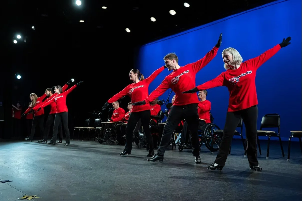 Group of people performing on stage, wearing matching red shirts, with some in wheelchairs, demonstrating a dance or movement routine.