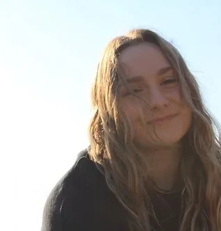 A young woman outdoors with long wavy blonde hair, smiling with her eyes closed, sunlight on her face, wearing a black top.
