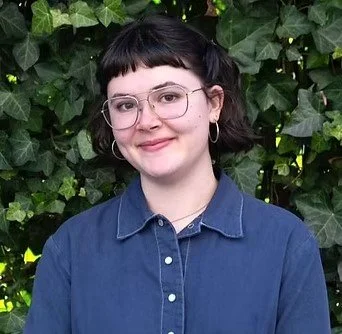 Portrait of a young woman with glasses and dark hair styled in pigtails, wearing a blue shirt, standing in front of green leafy plants.