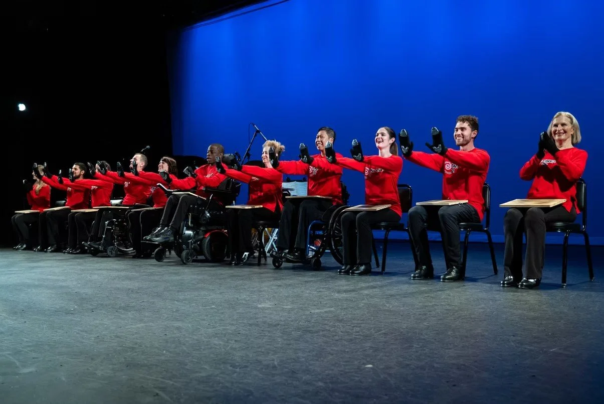 Group of performers with disabilities sitting on stage, extending their arms in a synchronized gesture, all wearing matching red shirts, with a dark background and blue lighting overhead.