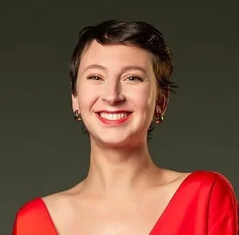 A woman with short brown hair, smiling, wearing a red dress and earrings against a dark background.