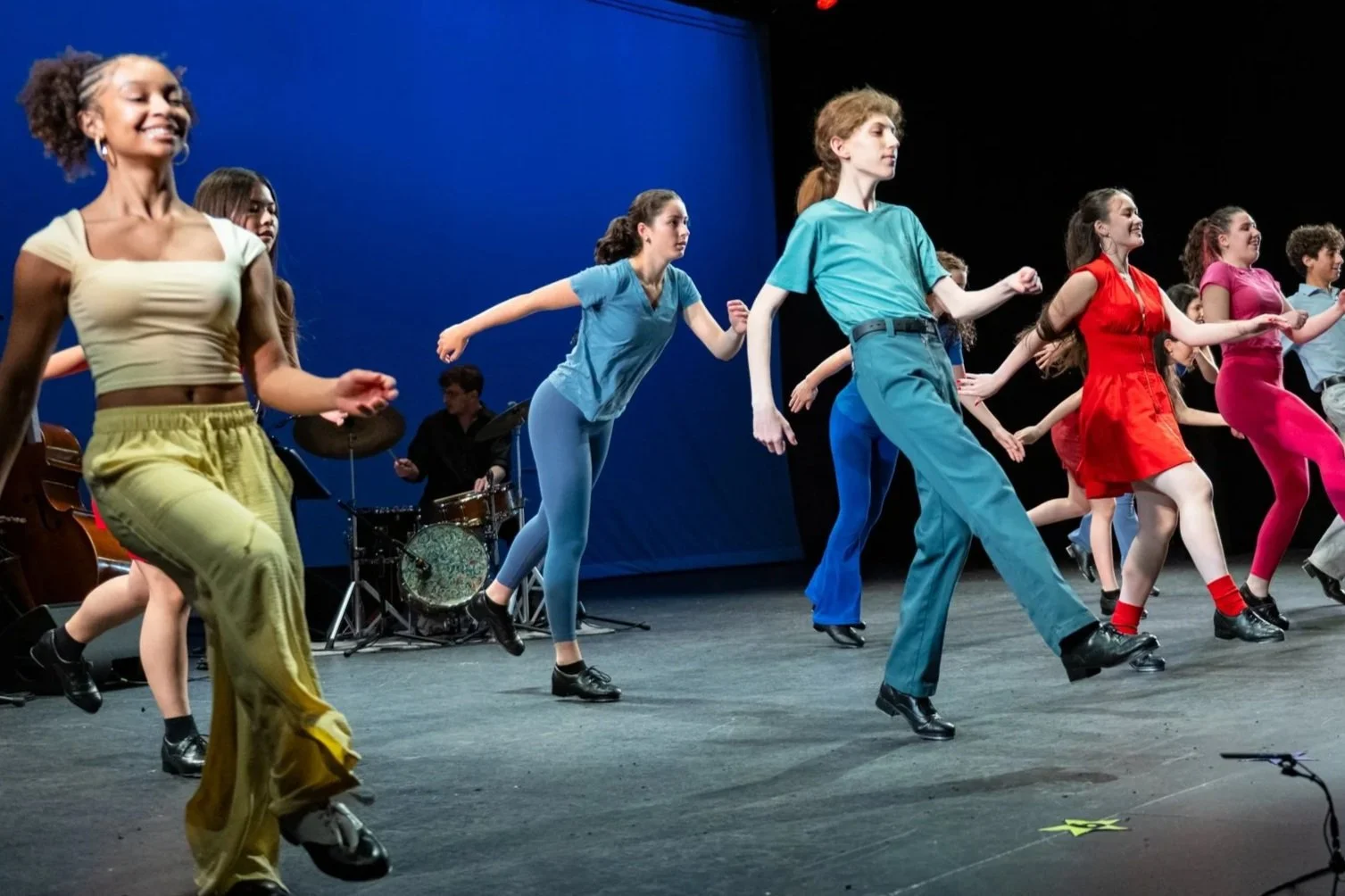Group of young dancers, dressed in colorful clothing, performing a dance on stage with a live band playing in the background.