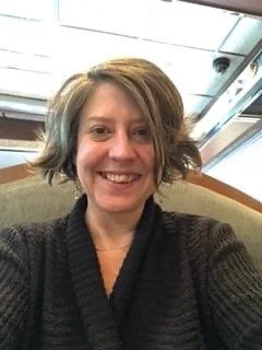 A woman smiling in an indoor setting with a ceiling and part of a chair visible in the background.