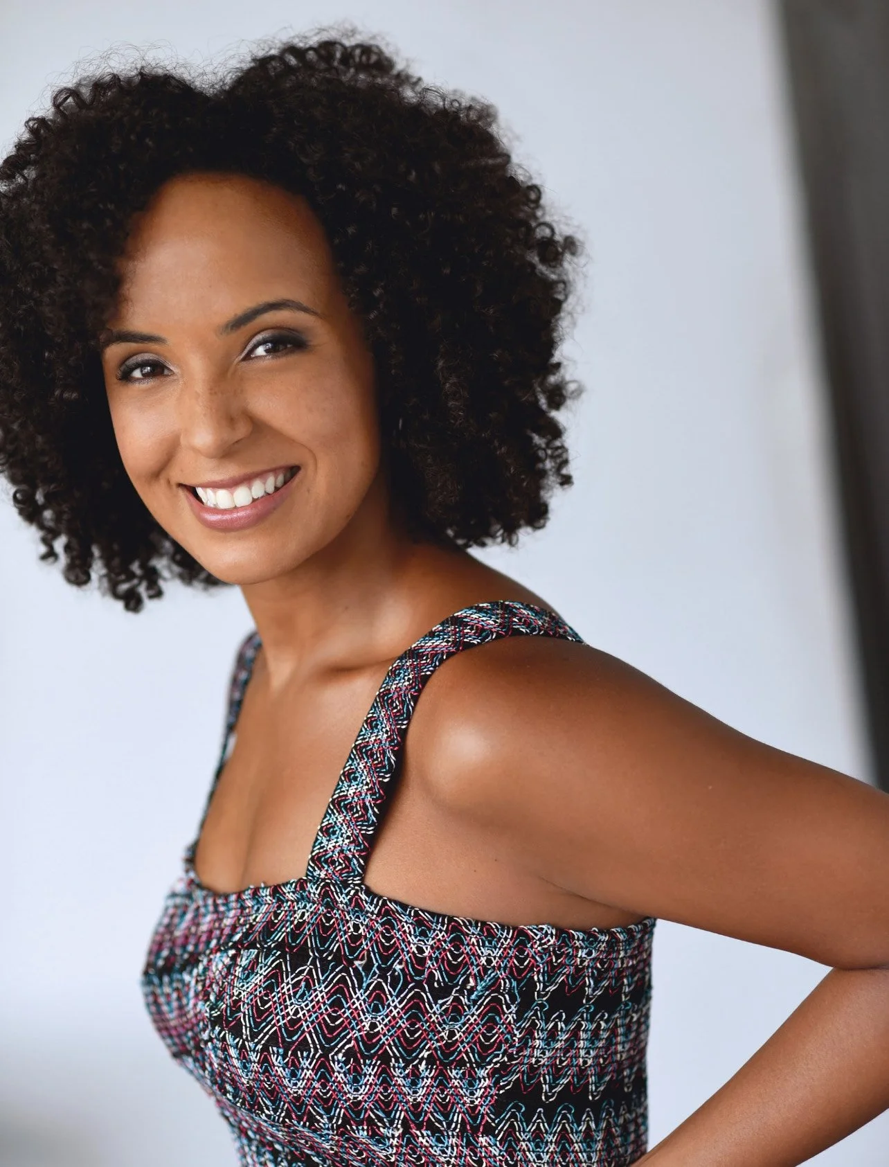 A woman with dark curly hair smiling at the camera wearing a patterned sleeveless top.