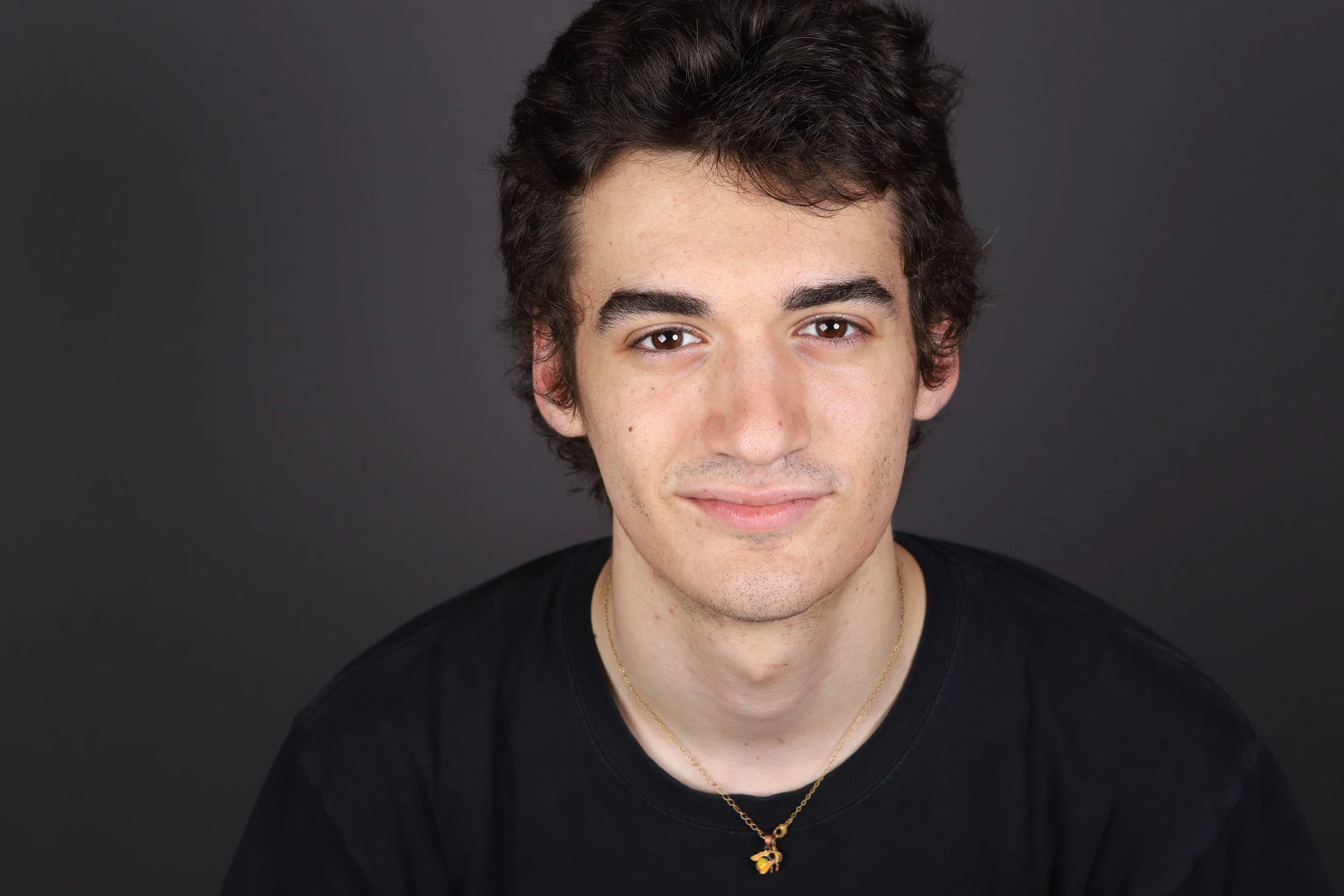 A young man with dark, wavy hair and light skin, wearing a black shirt and a gold necklace with a bee pendant, posing against a dark gray background.