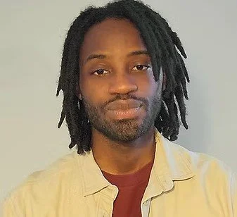A man with dreadlocks and a beard takes a selfie against a plain background, wearing a red shirt and a beige jacket.