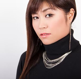 A woman wearing a black turtleneck with layered necklaces and a long earring, with long dark hair and a neutral expression.