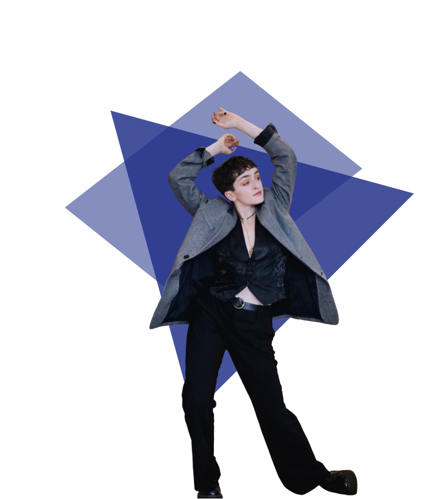 A young woman with short dark hair wearing a gray coat, black blouse, and black pants striking a pose with arms raised against a blue geometric background.