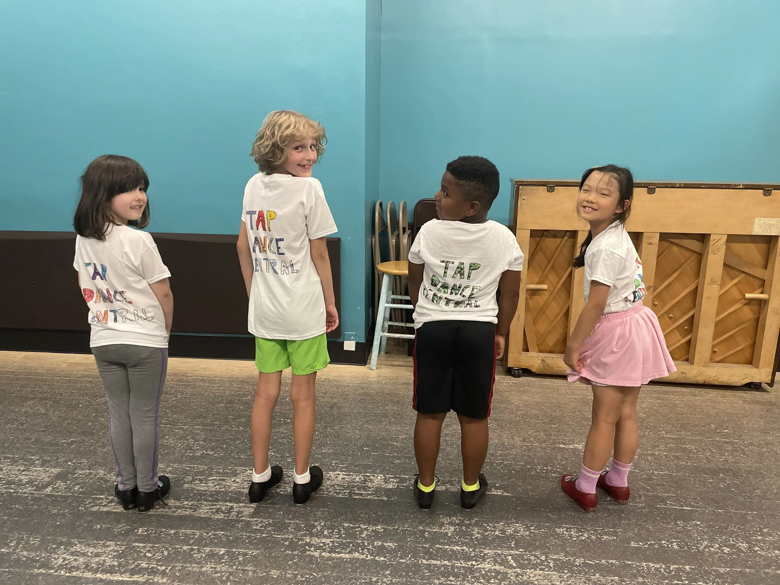 Four children standing on a dance floor, facing away from the camera, in a dance studio with teal walls and stacked chairs, wearing casual dance clothes.