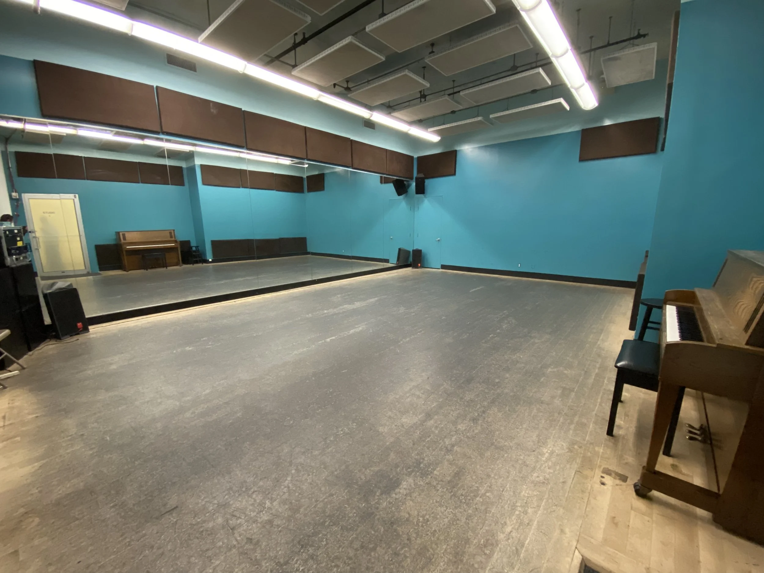 An empty dance studio with a large wall mirror, hardwood floors, blue walls, and two pianos, one on each side of the room, with chairs beside them.