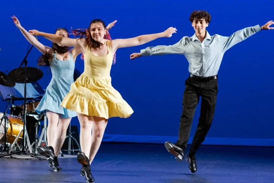 Two girls in colorful dresses and a boy in a white shirt and black pants performing a dance on stage with a blue background and musical instruments behind them.