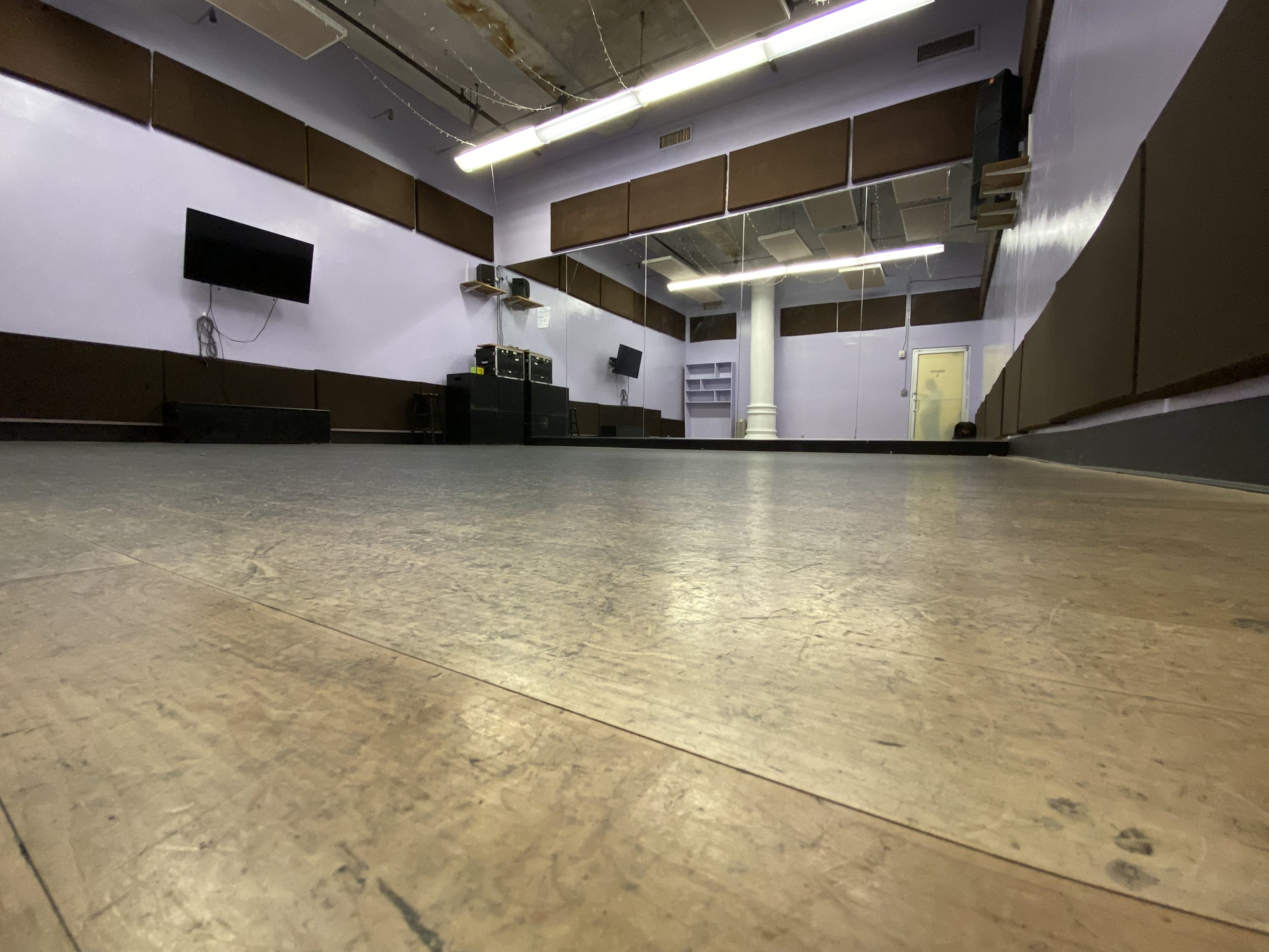 Empty dance studio with wooden floor, purple walls, and mirrors on the sides. Wall-mounted TV screens and audio equipment are visible.