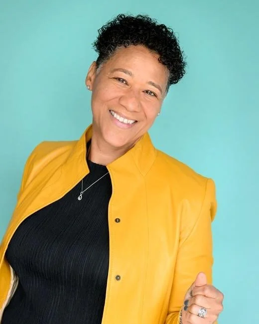 Woman smiling in a yellow jacket and black top against a light blue background.