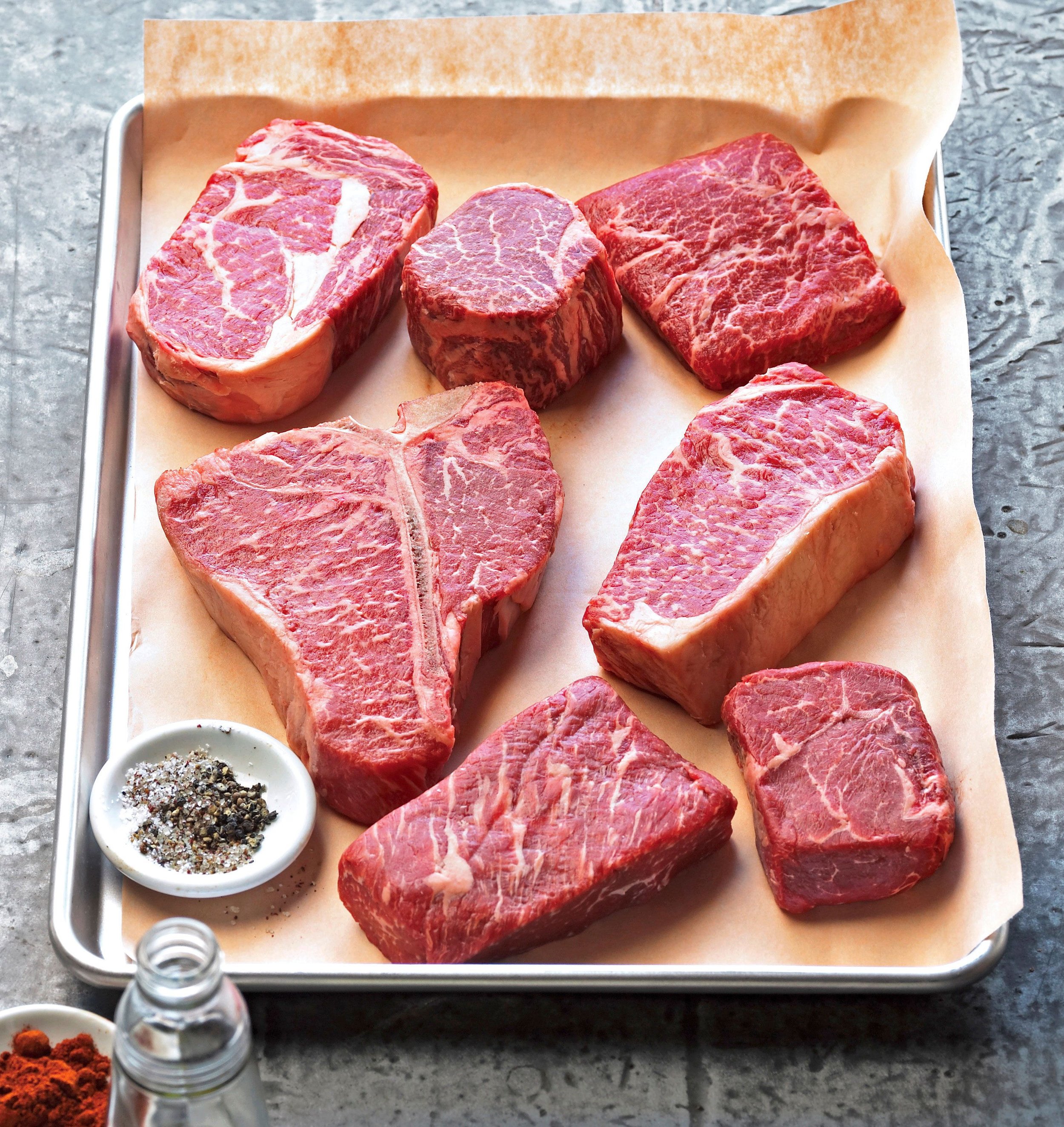 Assorted raw beef steaks on a baking sheet with seasoning and spices nearby.