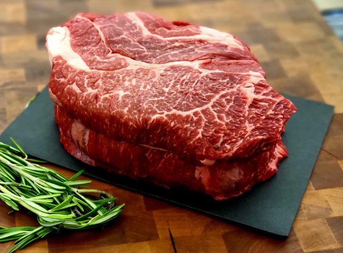 Raw marbled beef steak topped on a black cutting board with a sprig of rosemary beside it on a wooden surface.