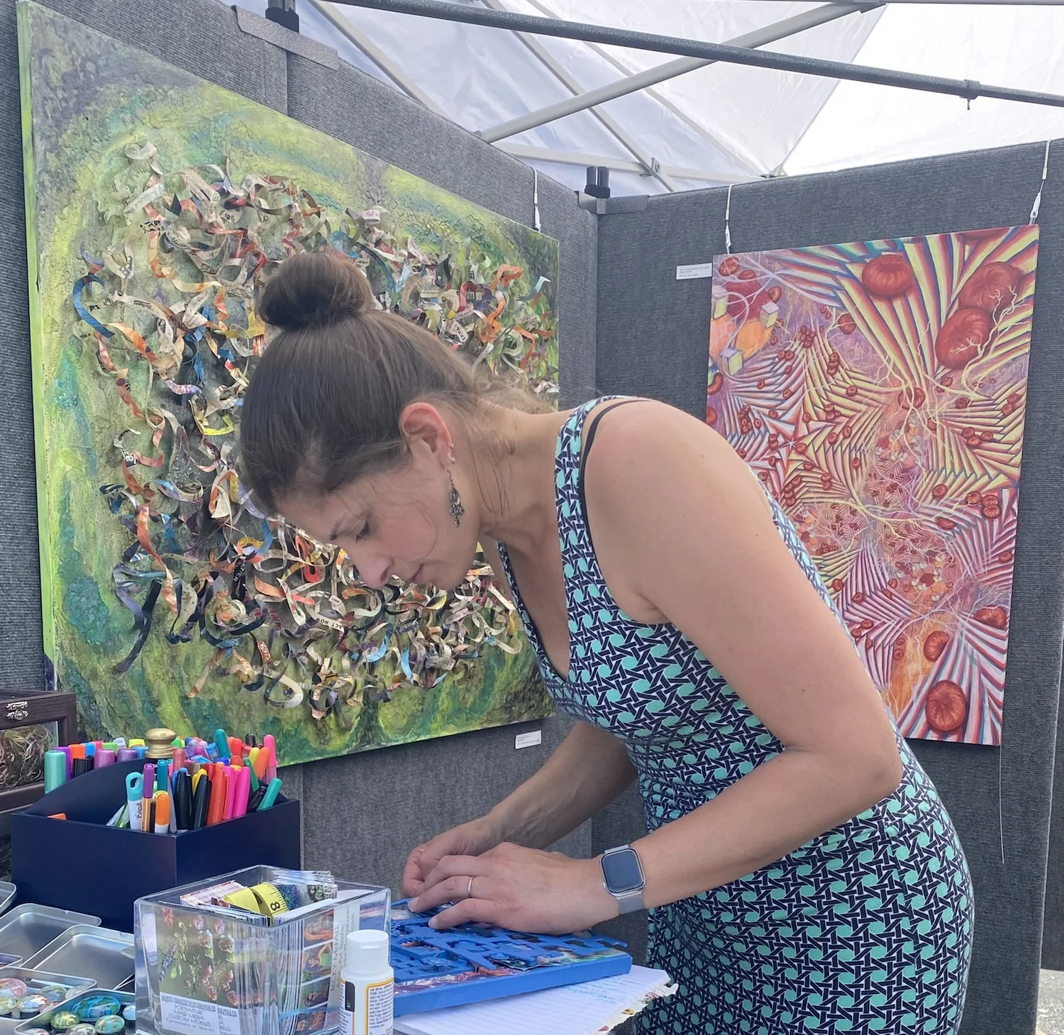 A woman with a bun hairstyle, wearing a patterned dress and a smartwatch, is setting up art supplies at an art booth. Behind her, two colorful paintings are displayed on a gray wall, one featuring twisted ribbons and the other depicting an abstract scene with red elements and streaked patterns.