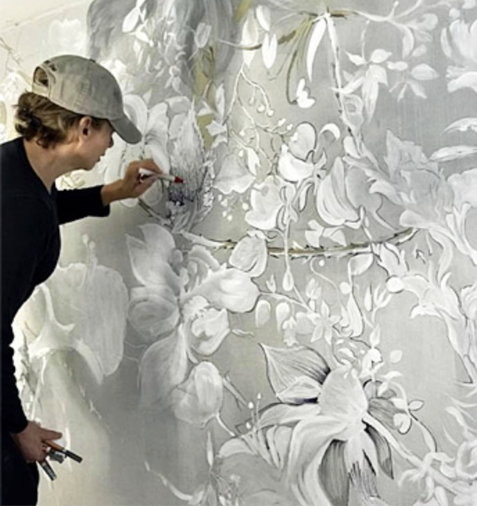 A person painting a large floral mural in shades of gray and white on a wall.