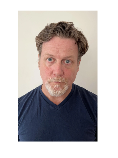 A man with curly brown hair, blue eyes, and a goatee, wearing a dark blue V-neck shirt, standing against a plain white background.