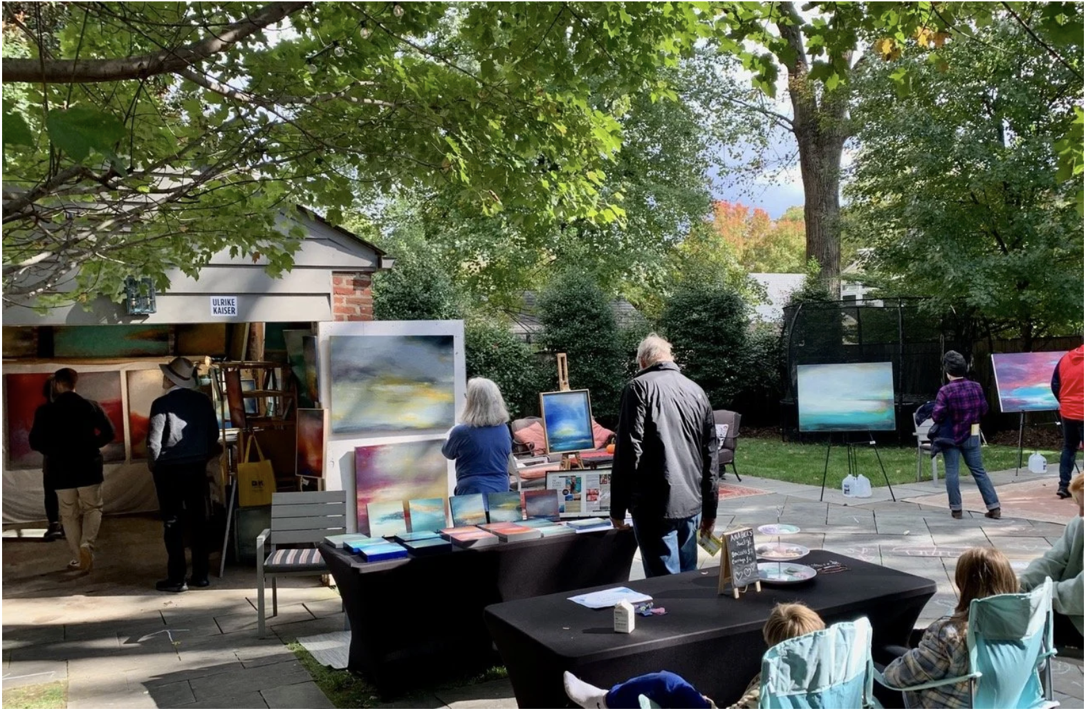 Outdoor art exhibit with people viewing landscape paintings displayed on easels and tables, trees, and a small building in the background.