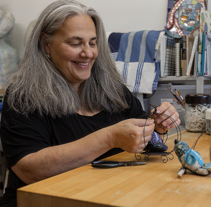 A woman with long gray hair smiling while working on an artistic project at a wooden table, surrounded by art supplies and colorful artwork in the background.