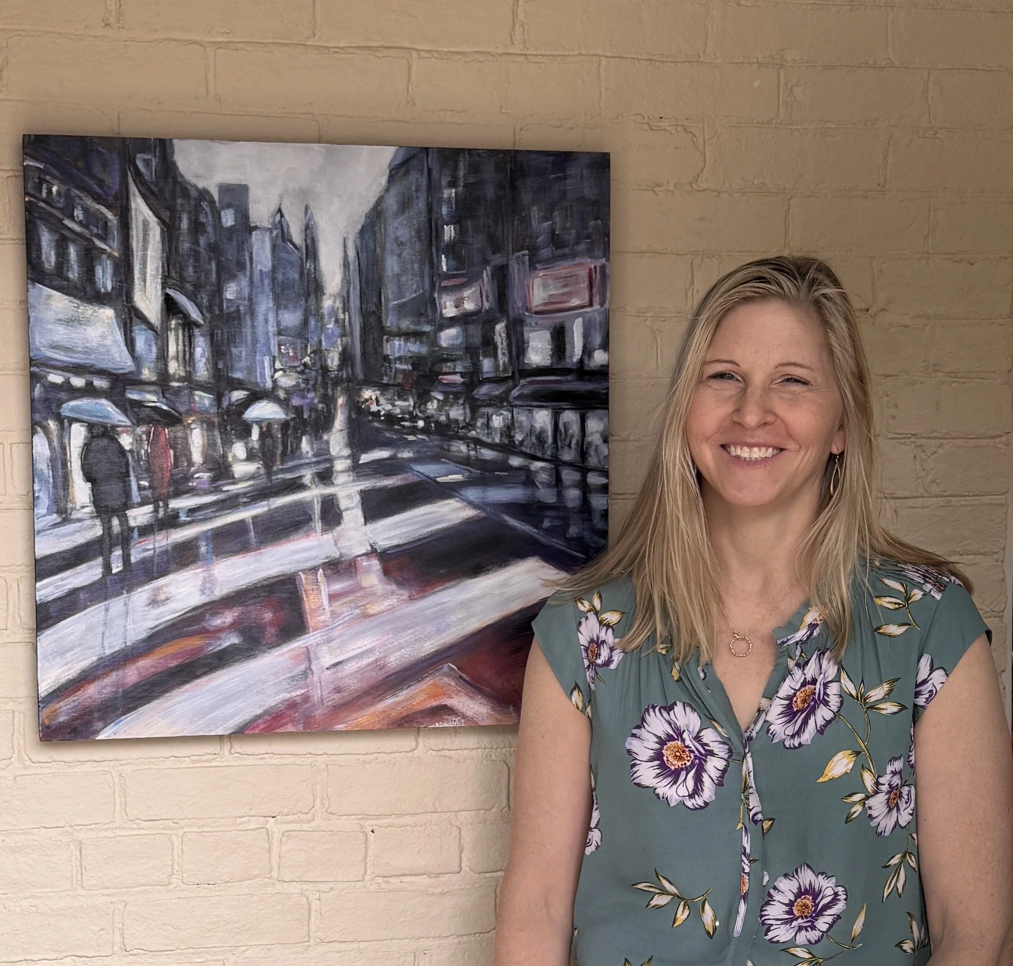A woman with blonde hair smiling, wearing a floral dress with purple flowers, standing in front of a painting of an urban street scene with cars and umbrellas, on a brick wall background.