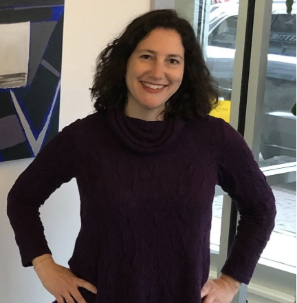 A woman with curly dark hair smiling, wearing a purple sweater, standing indoors near large glass windows.