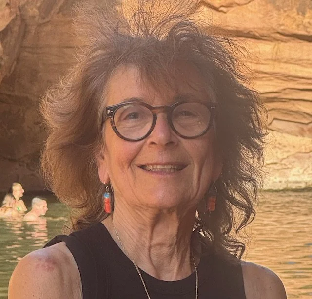 A smiling elderly woman with glasses and earrings relaxing outdoors near water with rocky cliffs in the background.