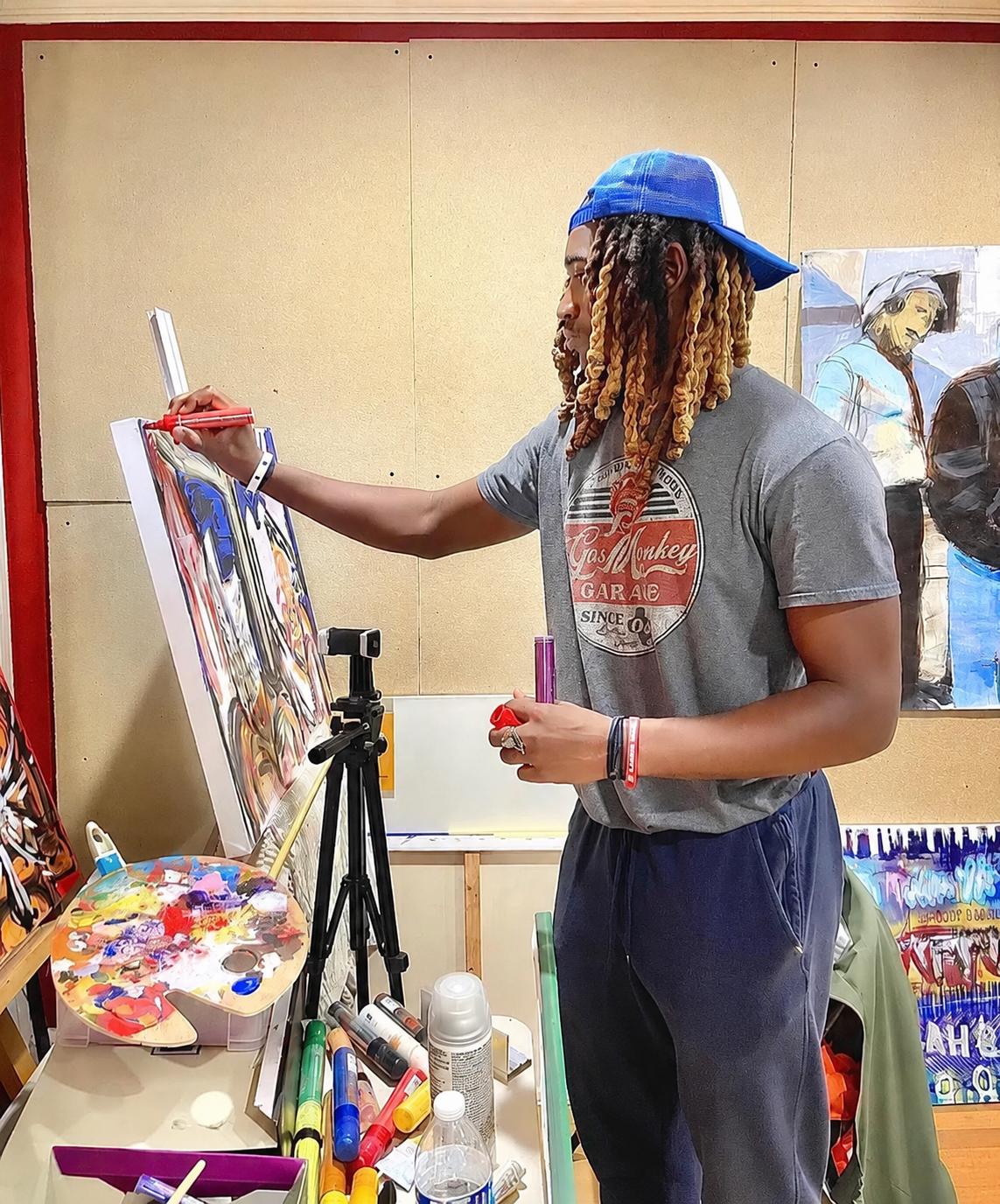 A man with long, purple-colored dreadlocks, wearing a gray T-shirt with a logo, a blue and white cap worn backwards, and dark sweatpants, is painting on a canvas set up on an easel in an art studio. He is holding a red marker or paintbrush and a pink marker. The table in front has various art supplies including paint markers, a paint palette, and bottles of spray paint, with a tripod camera positioned in front of him. In the background, there are paintings on the wall.