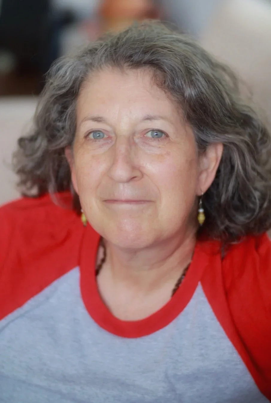 A middle-aged woman with curly gray hair and blue eyes wearing a red and gray shirt, and yellow earrings.