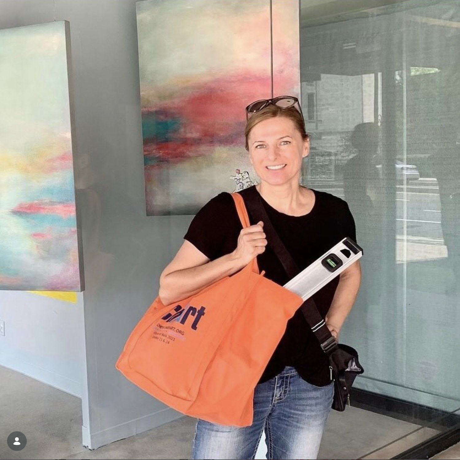 A woman with short brown hair, wearing a black shirt and blue jeans, standing in front of a glass wall with abstract artwork. She is smiling, holding an orange tote bag with a level tool and a black shoulder bag, and has sunglasses resting on her head.