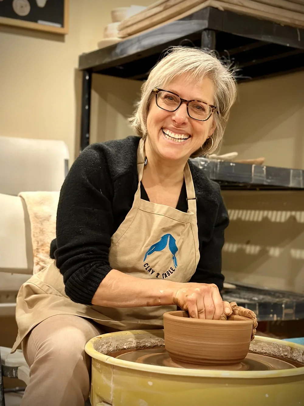 A smiling woman with glasses working on a pottery wheel, shaping a clay bowl in a pottery studio.