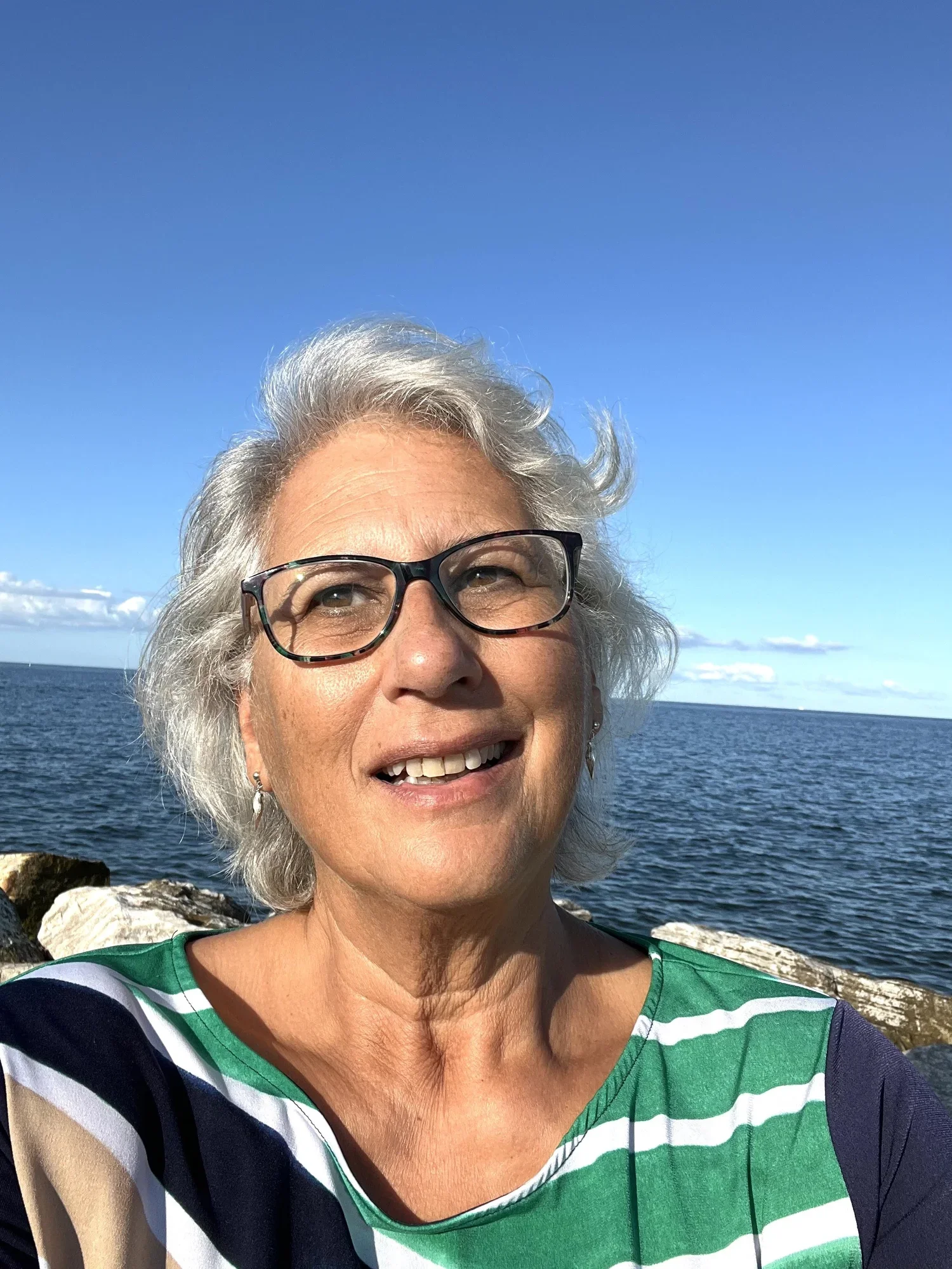 Close-up of a smiling woman with gray hair and glasses, wearing a green and white striped top, standing outdoors near water with rocks and a partly cloudy sky in the background.